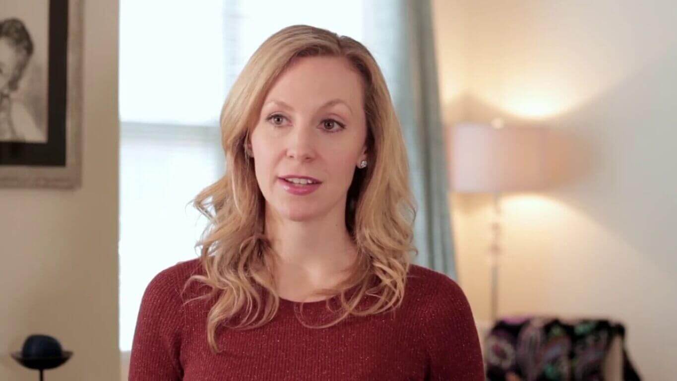 Blonde woman in a red sweater speaking indoors, with a lamp and framed picture in the background, natural light from window.